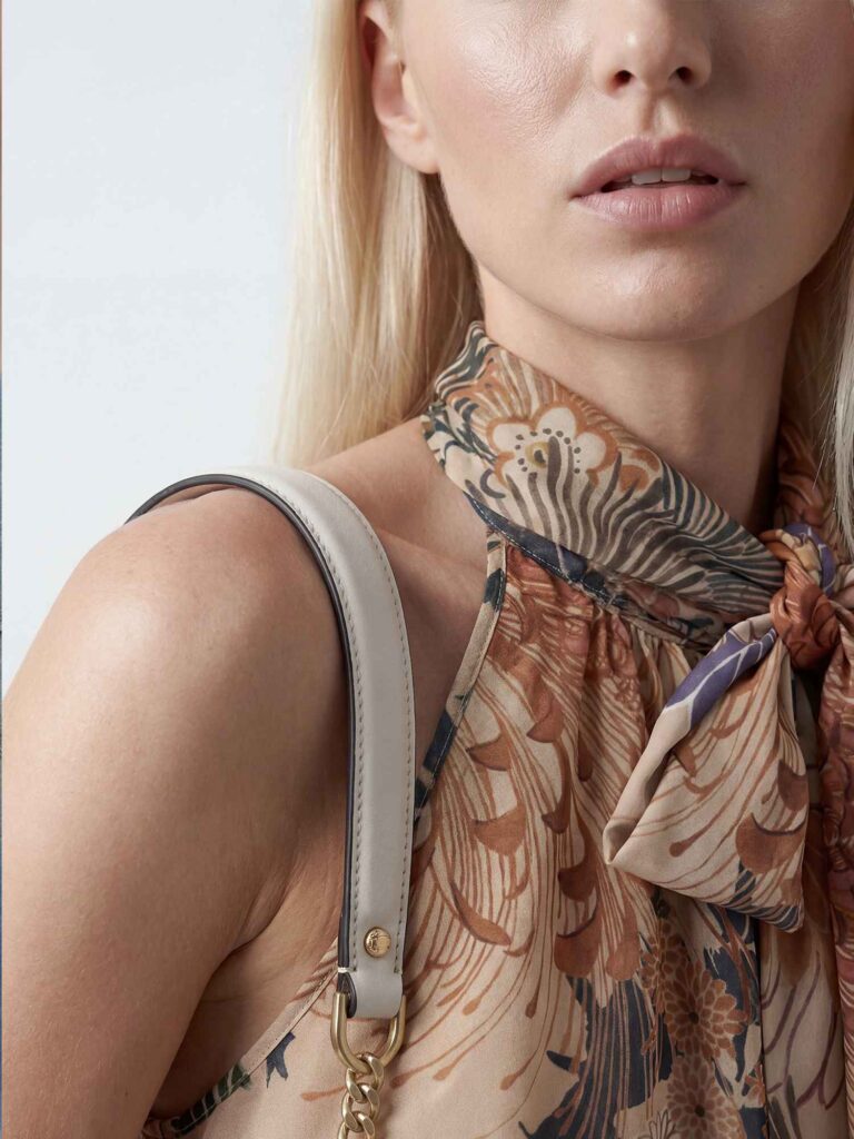 Primo piano parziale di una modella bionda con top floreale annodato al collo nei toni beige e rame. Si vedono labbra, collo, spalla e tracolla di una borsa chiara. Sfondo neutro chiaro.