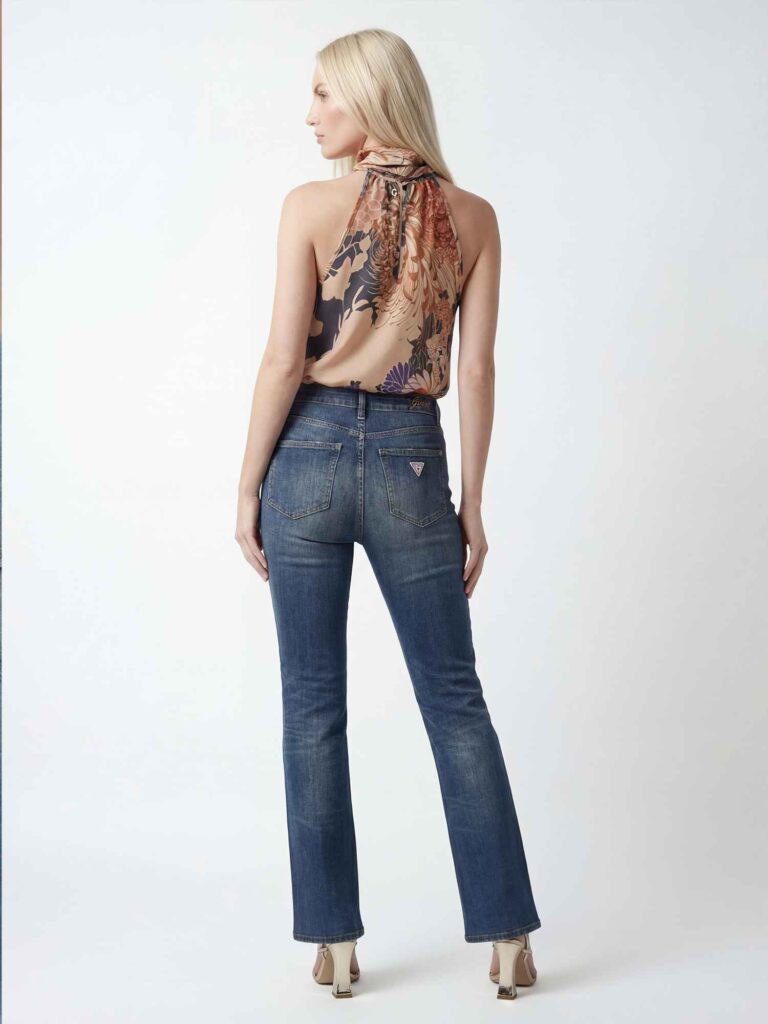 Vista posteriore a figura intera di una modella bionda che indossa top con schiena scoperta e jeans blu aderenti. La modella è in posa laterale su sfondo chiaro minimal.