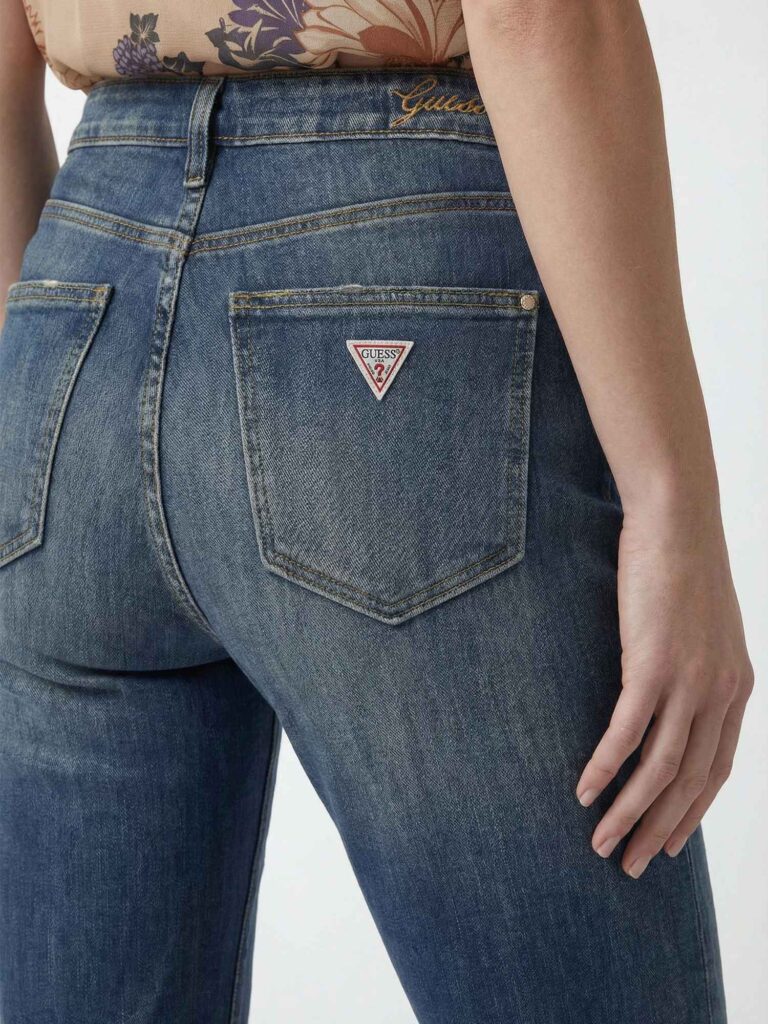 Dettaglio ravvicinato del retro di jeans blu da donna con tasche posteriori, cuciture a contrasto e logo triangolare GUESS applicato sulla tasca destra. Si vede anche parte del top floreale e del braccio della modella.