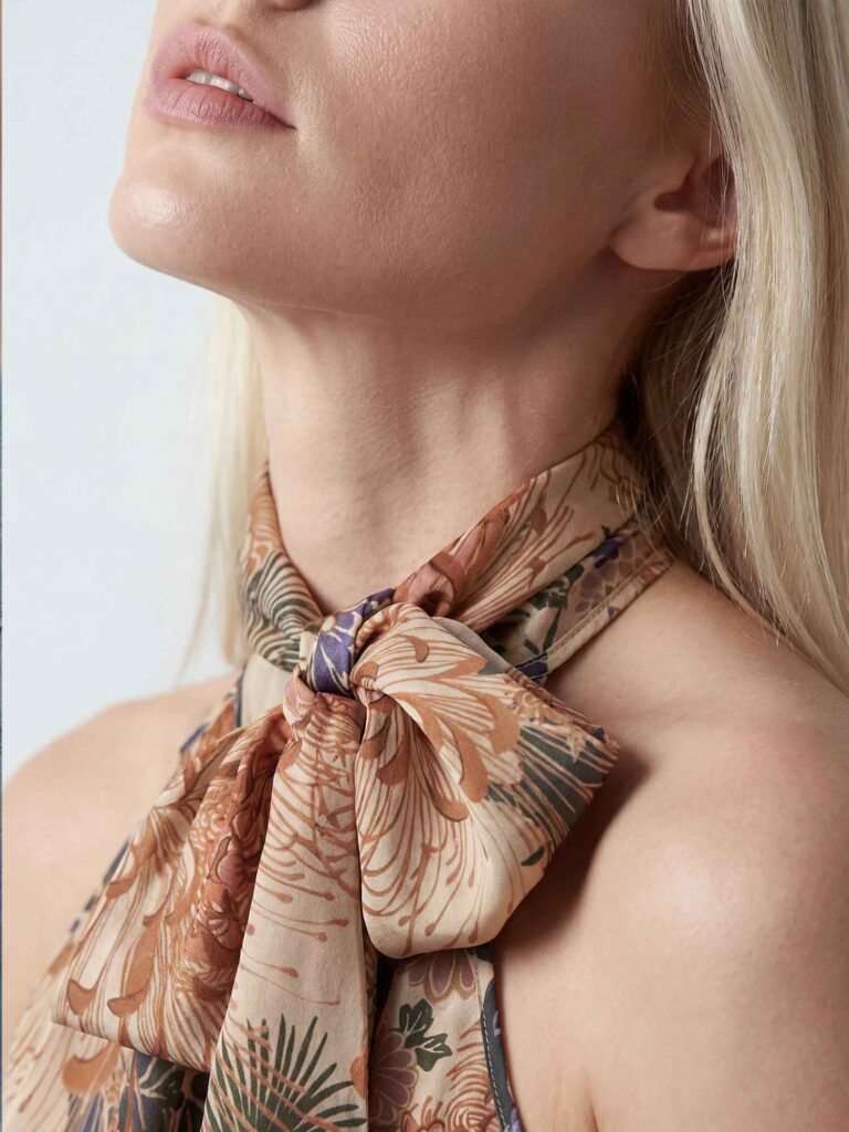 Dettaglio ravvicinato del collo e della spalla di una modella bionda con top floreale annodato al collo. Si vedono parte del viso, labbra e fiocco del tessuto nei toni beige, rame e blu.
