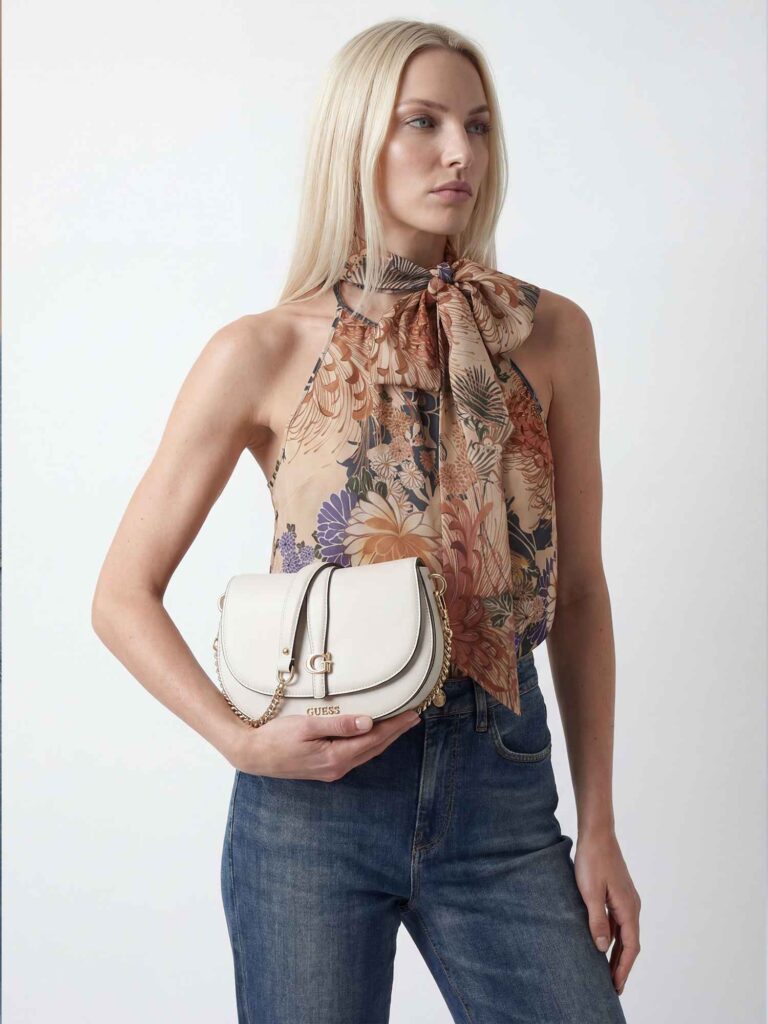 Vista a mezzo busto di una modella bionda che guarda lateralmente. Indossa top floreale e jeans blu, tenendo una borsa a tracolla bianca con dettagli metallici e logo GUESS frontale.
