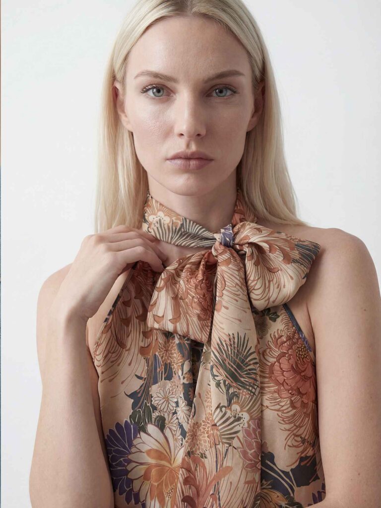 Primo piano frontale di una modella bionda che guarda in camera. Indossa top floreale con grande fiocco annodato al collo nei toni beige, rame e blu. Sfondo chiaro minimal.