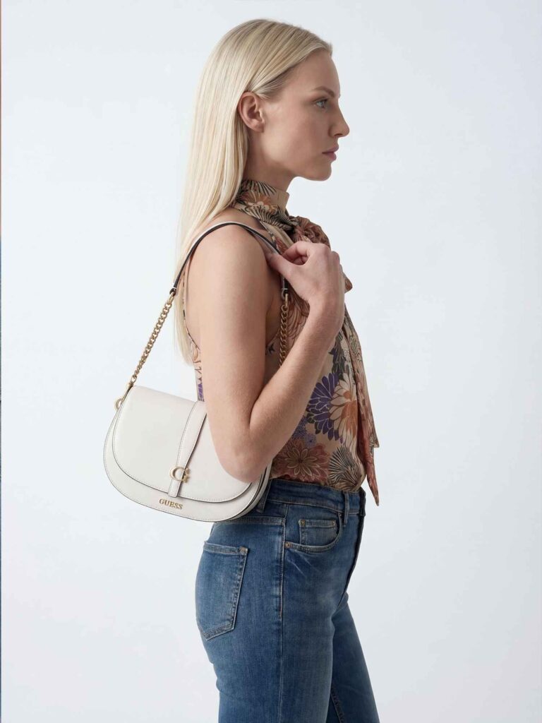 Vista laterale di una modella bionda con top floreale e jeans blu aderenti. Porta una borsa a spalla bianca con catena dorata e tiene la tracolla con una mano. Sfondo neutro chiaro.