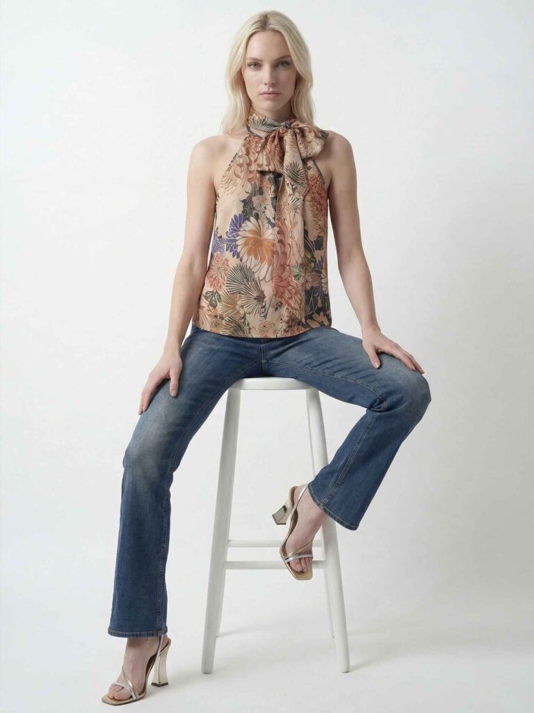 Vista frontale di una modella bionda seduta su uno sgabello bianco. Indossa top floreale, jeans blu e sandali con tacco, con posa elegante su sfondo chiaro essenziale.