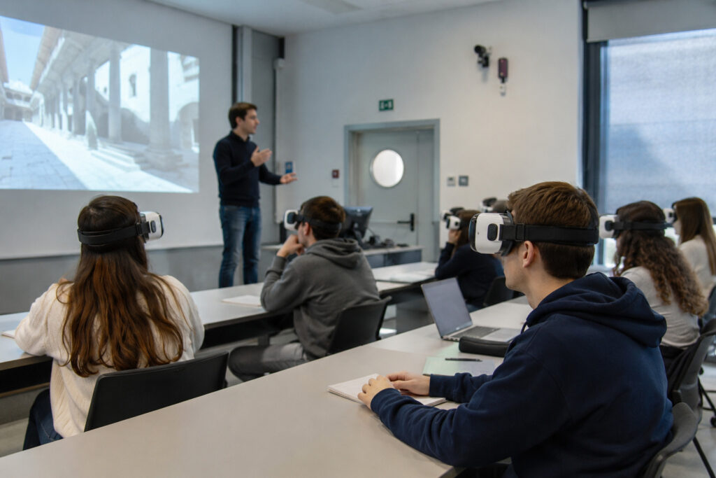 Aula universitaria moderna e luminosa con studenti seduti ai banchi che indossano visori VR durante una lezione. Un docente è in piedi davanti alla classe mentre spiega e gesticola, con un’immagine proiettata su uno schermo alle sue spalle. Gli studenti sono concentrati e alcuni hanno anche laptop e quaderni aperti sui tavoli.